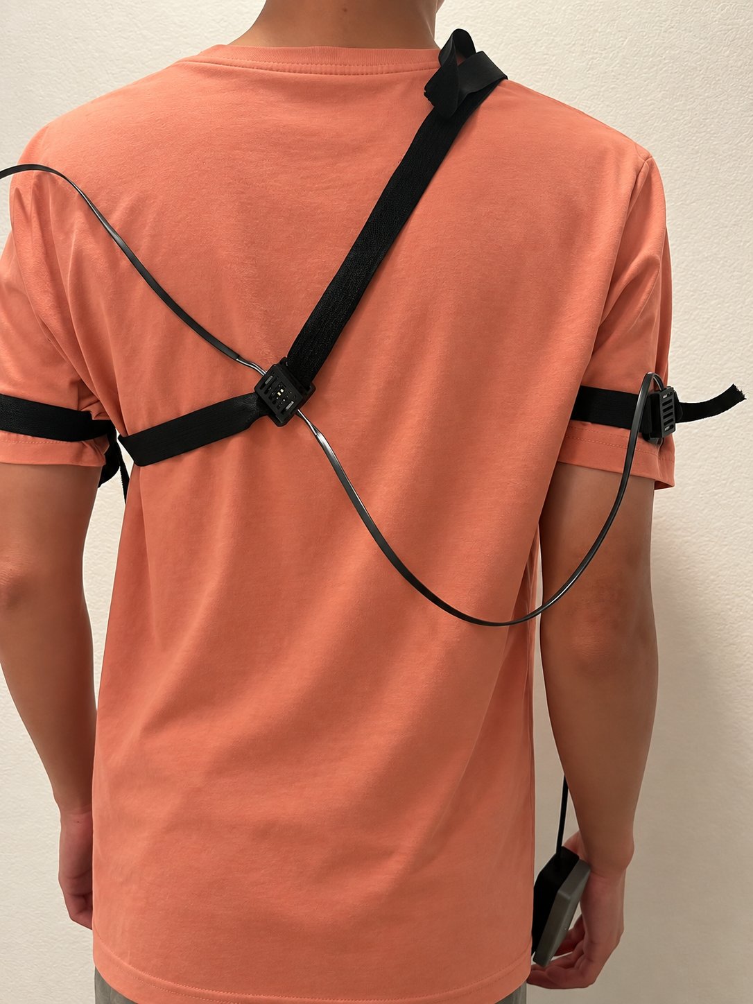 A person seen from behind wearing a shoulder-and-waist strap harness, with a cable running to a small handheld device at the waist